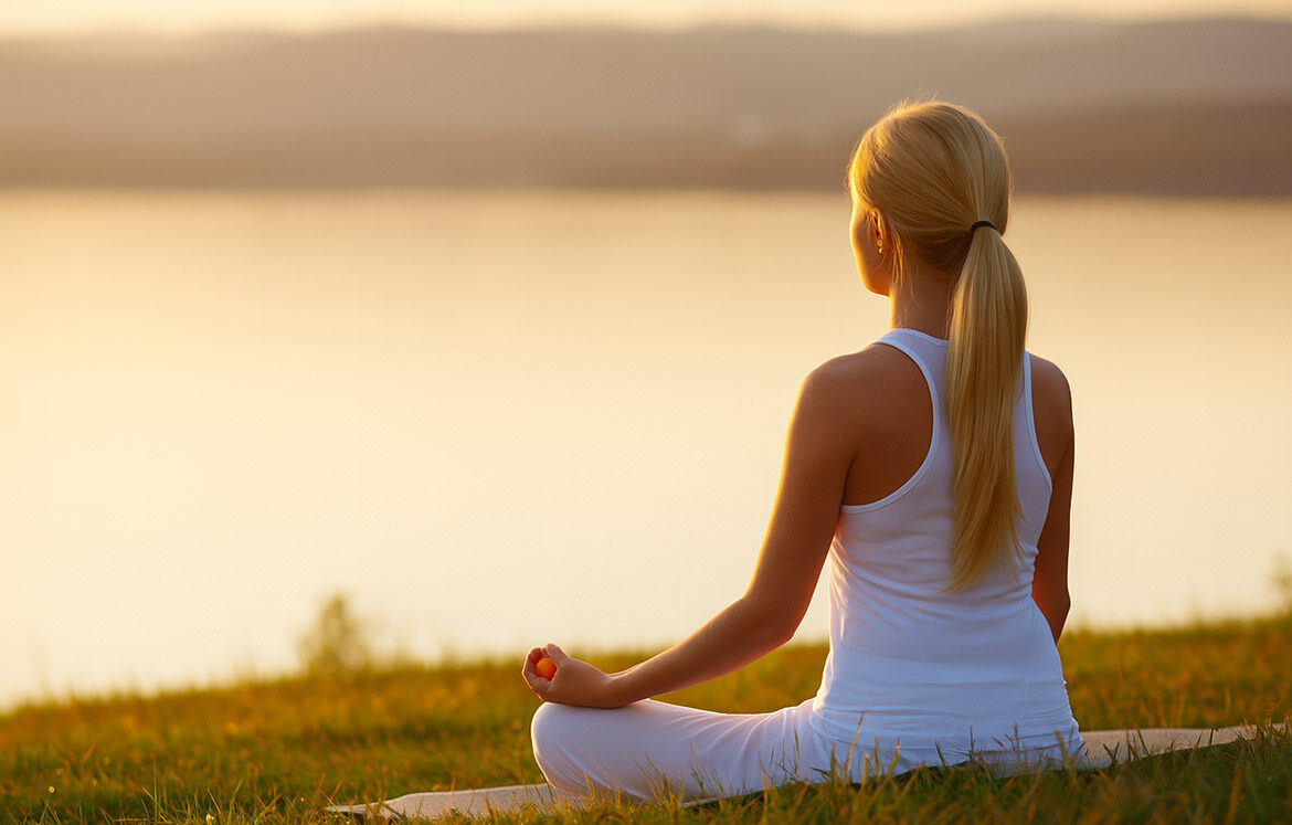meditacija Ženska sedi v jogijskem položaju ob jezeru med sončnim zahodom in meditira – simbolično postavljanje mej za notranji mir in ravnovesje.